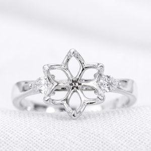 Puede incluir: Un anillo de tono plateado con un engaste central en forma de flor de seis pétalos diseñado para una piedra preciosa, flanqueado por dos pequeñas piedras claras en forma de pera a cada lado de la banda, con un acabado metálico pulido.