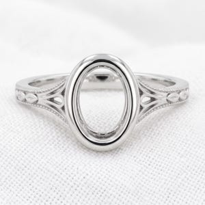 May include: A silver-toned metal ring featuring a central oval-shaped setting designed to hold a gemstone. The band displays intricate decorative detailing along the sides, including small leaf-like motifs and beaded edges surrounding the oval.