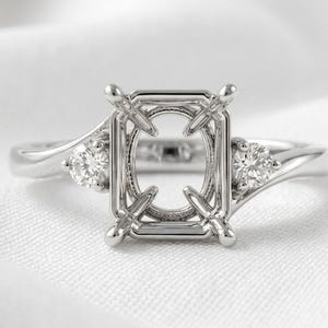 May include: A silver-toned metal ring setting featuring a central rectangular prong mount designed for a gemstone. The band includes a bypass-style design with a single round brilliant-cut clear stone set on each side of the central mounting.