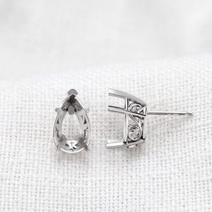May include: A pair of silver-toned metal earring settings designed for pear-shaped stones. Each setting features an open teardrop frame with intricate scrollwork detailing on the sides and a straight post extending from the back for attachment.