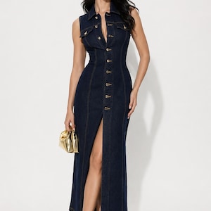 May include: Sleeveless, dark wash denim dress with a collared neckline and a button-down front. The dress features a high slit and is paired with gold-colored heels and a small gold handbag. The dress has a fitted silhouette.