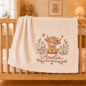 May include: A cream-colored knit baby blanket featuring an embroidered design of a fluffy highland cow surrounded by colorful wildflowers, a butterfly, and a bee. The name Amelia is stitched in pink script above the text Little Blessing.