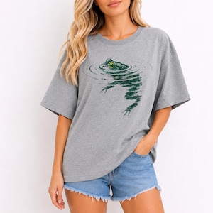 May include: A heather gray short-sleeve t-shirt featuring a dark green graphic print of a frog partially submerged in water, with ripples extending outward from the frog's head and body, creating a textured, artistic effect on the front.