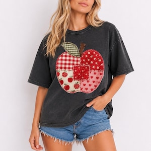 Op de afbeelding: Een antracietgrijs T-shirt met korte mouwen en een grote, centrale appelvormige afbeelding. De appel is samengesteld uit verschillende stoffen in patchworkstijl, waaronder rood-wit gingham, bloemenprints en kleine rode hartjes en appeltjes.