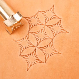 Könnte beinhalten: Ein metallenes Lederstempelwerkzeug mit einer sternförmigen Spitze neben einer hellbraunen Lederoberfläche. Das Leder weist ein sich wiederholendes geometrisches Muster aus eingeprägten Sternformen auf, die durch das Werkzeug entstanden sind.