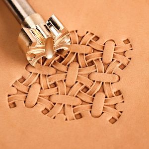 May include: A metal leather stamping tool with a cylindrical shaft and a complex, multi-pronged head is pressed into a piece of light tan leather, creating a repeating, woven basket-weave pattern with deep, embossed geometric impressions.