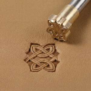 May include: A leather embossing tool featuring a Celtic knot design. The metal stamp is pressed into the tan leather, creating an embossed pattern. The tool has a brass head and a silver handle.
