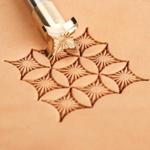 May include: A metal leather stamping tool with a starburst pattern head is pressed into a piece of light tan leather, creating a repeating geometric design of embossed starbursts and smooth almond-shaped petals in a square grid formation.