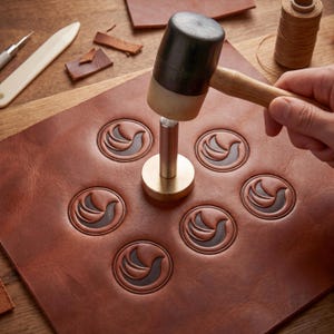 Peut inclure: Artisanat du cuir en cours. Une pièce de cuir marron avec des motifs d'oiseaux en relief est travaillée avec un maillet et un tampon métallique. D'autres outils et des chutes de cuir sont visibles sur une surface en bois.
