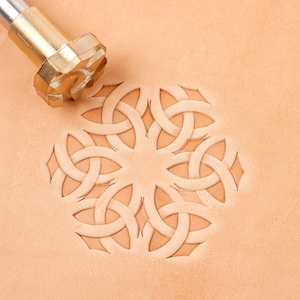May include: A brass leather stamping tool with a cylindrical handle and a circular head featuring a complex, repeating geometric knot pattern. The tool is positioned above a light tan leather surface that displays a deep, crisp impression of the design.