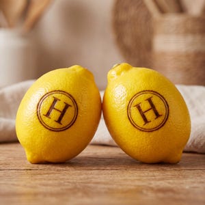 May include: Two bright yellow lemons placed side by side, each featuring a dark brown circular stamp with the capital letter H centered inside. The lemons have a textured, porous peel and are positioned upright on a wooden surface.