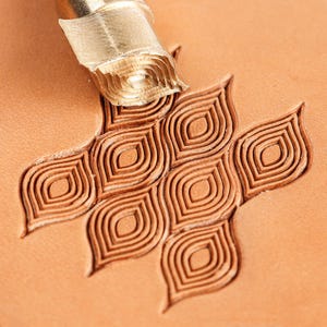 May include: A metal leather stamping tool positioned at the top of a tan leather surface, showing a repeating pattern of embossed, almond-shaped motifs with concentric lines pressed into the material by the tool's textured, pointed head.