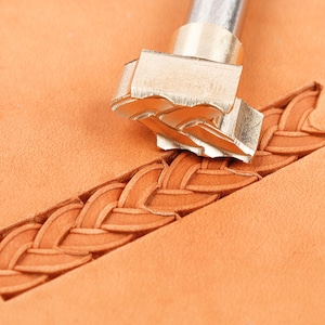Op de afbeelding: Een metalen lederstempelgereedschap met een gevlochten patroonkop, geplaatst op een stuk bruin leer. Het gereedschap heeft een cilindrische schacht en een gedetailleerde stempel die een doorlopende geweven vlechtindruk in het leer achterlaat.