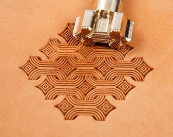 May include: A metal leather stamping tool with a cylindrical shaft and a patterned head is pressed into a piece of light tan leather, creating a repeating geometric design of interlocking lines and small diamond shapes in the surface of the material.