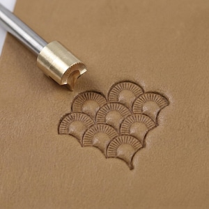May include: A leather craft tool with a brass head and a metal handle, positioned above a stamped pattern of fan-shaped designs on tan leather. The tool is designed for leatherworking and creating decorative patterns.