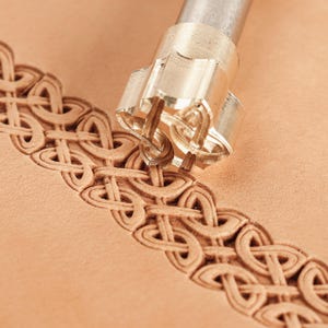 May include: A metal leather stamping tool with a cylindrical shaft and a patterned head is pressed into a piece of light tan leather, creating a continuous, repeating embossed design of interconnected loops and geometric shapes along the surface.