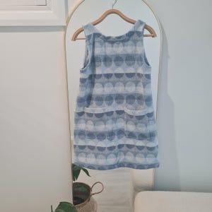 Peut inclure: Robe sans manches bleu clair avec un motif répétitif de grands cercles superposés dans des tons de bleu et de blanc. La robe a deux poches avant et est suspendue à un cintre en bois. La robe se reflète dans un grand miroir cintré.