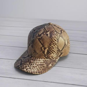 Peut inclure: Une casquette de baseball pr&eacute;sentant un motif en peau de serpent marron et beige sur toute la surface. La casquette poss&egrave;de une calotte structur&eacute;e et une visi&egrave;re incurv&eacute;e, avec un bouton au sommet et des &oelig;illets d'a&eacute;ration sur les panneaux.