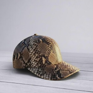 May include: A baseball cap featuring a brown, tan, and black snakeskin pattern. The cap has a structured crown and a curved brim, with the textured scale print covering the entire exterior surface of the headwear in a consistent design.