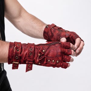 Può includere: Guanti senza dita con finitura rosso scuro invecchiato, caratterizzati da dettagli in metallo color ottone tra cui cinturini con fibbia, occhielli circolari, piccole borchie e una cerniera funzionale sul dorso della mano e sul polso.