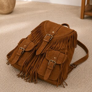 May include: A brown suede backpack featuring long fringe detailing across the front flap and two exterior pockets. The bag includes adjustable straps with metal buckles and metallic stud accents on the flap and pocket closures for secure fastening.