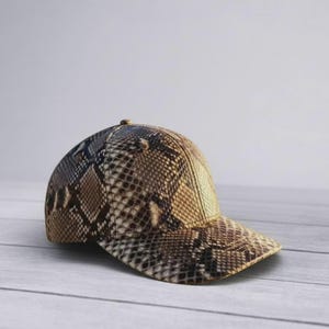 Peut inclure: Une casquette de baseball avec un motif peau de serpent dans des tons marron, beige et noir. La casquette pr&eacute;sente une calotte structur&eacute;e et une visi&egrave;re incurv&eacute;e, avec un imprim&eacute; &eacute;cailles textur&eacute; recouvrant toute la surface ext&eacute;rieure.