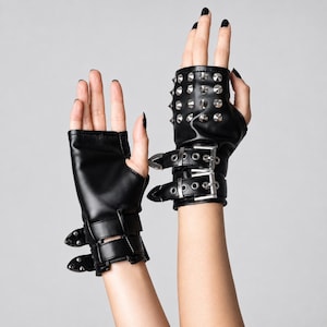 Könnte beinhalten: Ein Paar schwarze fingerlose Handschuhe aus glattem Material. Die Handschuhe verfügen über silberfarbene Metallnieten auf dem Handrücken und zwei verstellbare Riemen mit silberfarbenen Schnallen und Ösen am Handgelenk für einen sicheren Sitz.