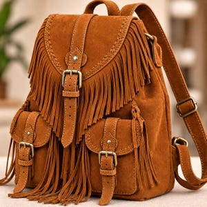 May include: A tobacco brown suede backpack featuring long fringe detailing on the front flap and two exterior pockets. The bag includes adjustable straps with antique brass-toned metal buckles and braided trim accents along the edges of the flap.