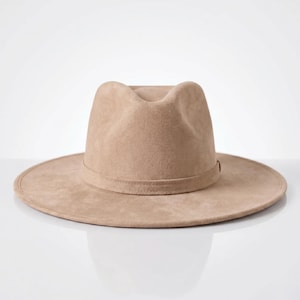 May include: A light tan fedora hat with a wide, flat brim and a pinched crown. The hat features a smooth, suede-like texture and a matching tonal band encircling the base of the crown, creating a clean, uniform appearance in a neutral earth tone.