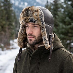 May include: A dark brown leather trapper hat featuring thick, multi-tonal brown and black faux fur lining on the ear flaps and forehead. The hat includes adjustable leather chin straps with silver-toned metal buckles and snap button closures.