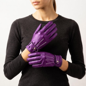May include: A pair of vibrant purple leather gloves featuring white contrast stitching along the fingers and back of the hand. Each glove includes a wrist strap with a metallic snap button closure, providing a secure fit and a distinct design detail.