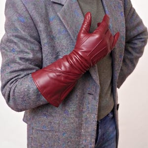 May include: A long, deep red leather glove with visible stitching details along the fingers and back of the hand, worn with a textured, multi-colored grey and blue blazer over a solid grey shirt and blue denim jeans.