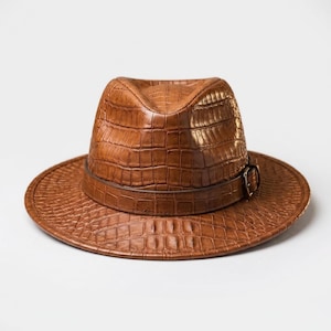 Könnte beinhalten: Ein hellbrauner Fedora-Hut mit einer strukturierten Krokodilleder-Optik auf der Krone und der breiten Krempe. Der Hut verfügt über ein passendes braunes Band um die Basis der Krone, das mit einer kleinen, dunklen Metallschnalle befestigt ist.