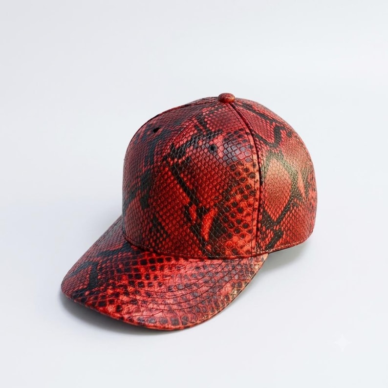 May include: A baseball cap featuring a vibrant red and black snakeskin pattern. The cap has a structured crown with a button at the top and a curved brim. The textured material mimics the appearance of reptile scales across the entire surface.