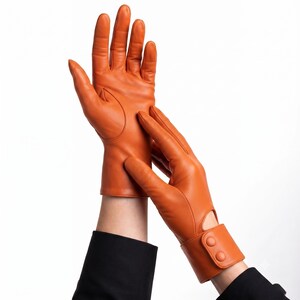 May include: A pair of vibrant orange leather gloves featuring a smooth finish, five fingers, and a wrist strap detail with two matching buttons. The gloves extend to the wrist and include a small cutout design near the cuff for a tailored fit.