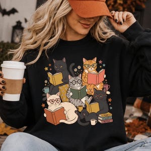May include: A black long-sleeve sweatshirt featuring a graphic print of five cats reading books. The cats are depicted in various colors including black, orange, and cream, wearing glasses and surrounded by small stars, flowers, and coffee mugs.