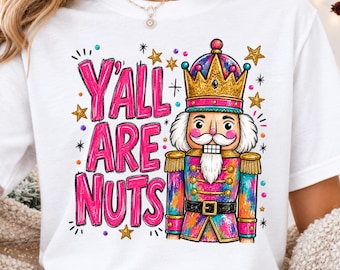 May include: A white short-sleeved t-shirt featuring a colorful graphic of a nutcracker wearing a gold crown. The text Y'ALL ARE NUTS is printed in large, bright pink letters to the left of the nutcracker, surrounded by small gold stars.