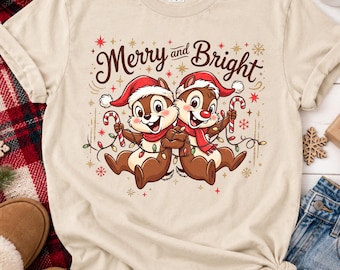 May include: A beige short-sleeve t-shirt featuring a graphic of two cartoon chipmunks wearing red Santa hats and scarves. They are holding candy canes and tangled in Christmas lights. The text Merry and Bright is printed above them.