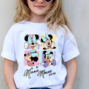 Pu&ograve; includere: T-shirt bianco con una grafica colorata di Minnie Mouse in quattro pose diverse, ciascuna in un quadrato. La maglietta ha le parole "Minnie Mouse xoxo" in corsivo. La ragazza indossa occhiali da sole a forma di cuore.
