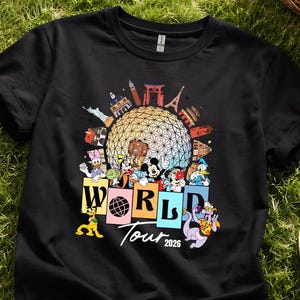 May include: Black t-shirt with a vibrant graphic of Disney characters and world landmarks. The design includes the text "WORLD Tour 2026" with characters such as Mickey Mouse and Donald Duck. The shirt is shown on a grassy background.