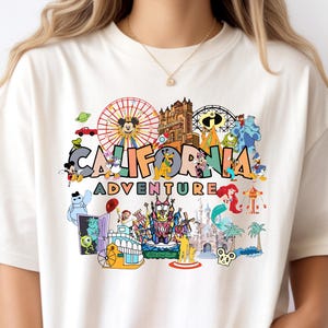 May include: A cream-coloured t-shirt with a vibrant graphic. The design features the words "CALIFORNIA ADVENTURE" alongside illustrations of a Ferris wheel, cartoon characters, and a castle, reminiscent of a theme park.