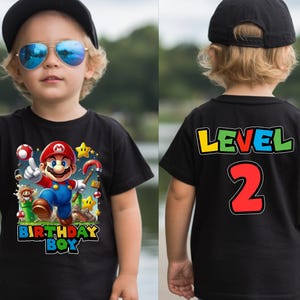 May include: Black t-shirt featuring a colorful graphic of Mario with the text "BIRTHDAY BOY" on the front. The back of the shirt displays "LEVEL 2". The shirt is worn by a child wearing a black cap and sunglasses.