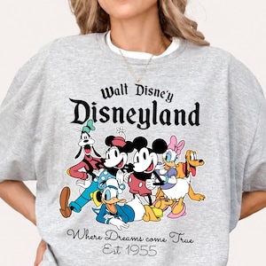 Pu&ograve; includere: Una felpa grigio chiaro con una grafica colorata dei personaggi Disney, tra cui Topolino, Minnie, Paperino e Pippo. Il testo "Walt Disney's Disneyland" e "Where Dreams come True Est. 1955" sono stampati sopra e sotto i personaggi.