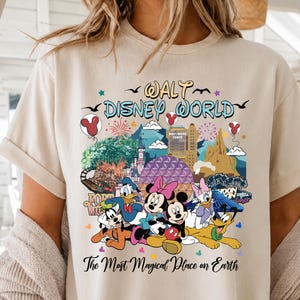 K&ouml;nnte beinhalten: Beiges T-Shirt mit einem farbenfrohen Aufdruck von Walt Disney World-Charakteren und -Sehensw&uuml;rdigkeiten. Das Design zeigt Micky und Minnie Maus, Donald Duck und Goofy, mit dem Text "Walt Disney World" und "The Most Magical Place on Earth".