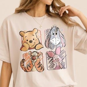 May include: Beige t-shirt featuring four cartoon character illustrations: Winnie the Pooh, Eeyore, Tigger, and Piglet. The characters are set within individual colored squares. The shirt is made of a soft, comfortable material.