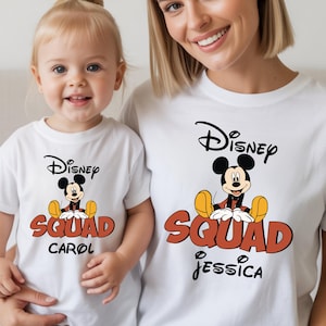 Op de afbeelding: Witte T-shirts met een Disney Mickey Mouse-ontwerp. De shirts tonen de woorden "Disney Squad" in oranje en zwart, met gepersonaliseerde namen eronder. E&eacute;n shirt heeft "Carol", de andere "Jessica".