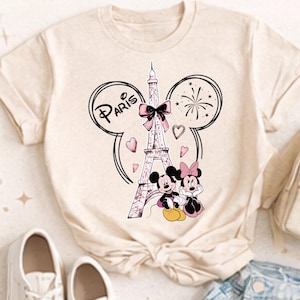K&ouml;nnte beinhalten: Beiges T-Shirt mit einem Aufdruck des Eiffelturms, Mickey und Minnie Mouse. Das Wort "Paris" steht &uuml;ber dem Turm in einem Mickey Mouse-Ohren-Design, mit Herzen und Feuerwerk. Die Figuren sind in Schwarz, Rosa und Gelb gehalten.