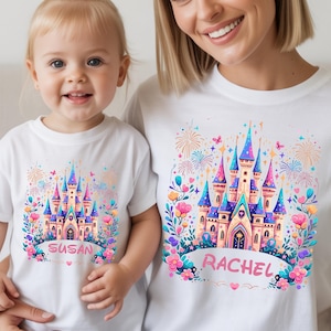 K&ouml;nnte beinhalten: Wei&szlig;e T-Shirts mit einem farbenfrohen Schlossmotiv. Das Schloss hat blaue, rosa und lila T&uuml;rme, umgeben von Blumen und Feuerwerk. Ein Shirt zeigt "SUSAN", das andere "RACHEL".