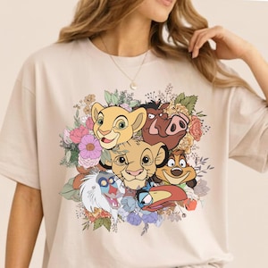 May include: Beige t-shirt featuring a colorful graphic of characters from The Lion King, including Simba, Nala, Pumbaa, Timon, and Rafiki, surrounded by a floral arrangement. The design is printed on a short-sleeved tee.