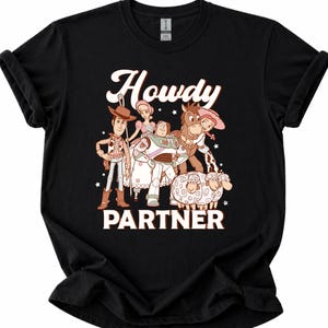 May include: Black t-shirt featuring a graphic of characters from a popular animated movie. The design includes the text "Howdy PARTNER" in white and brown. The shirt showcases a cowboy, space ranger, and other characters.